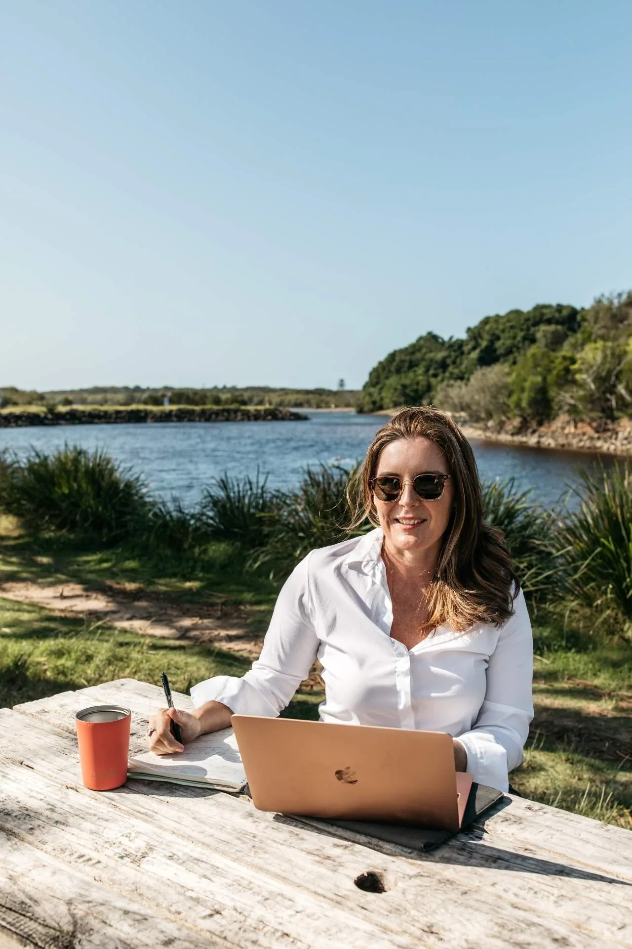 Michelle Williams working on a laptop at a picnic table outdoors, with a scenic ocean view behind her.