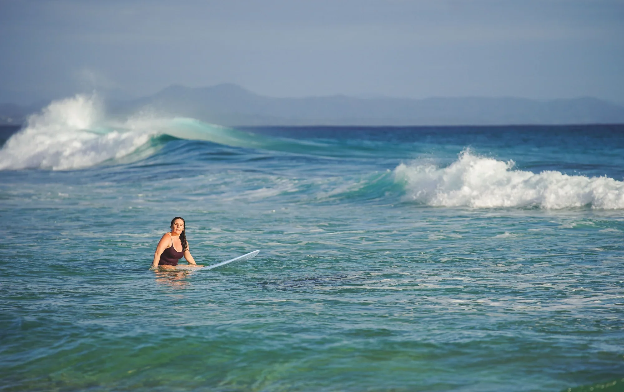 Michelle Williams paddling out on her surfboard at Byron Bay, back to camera, facing a large breaking wave with mountains in the distance.